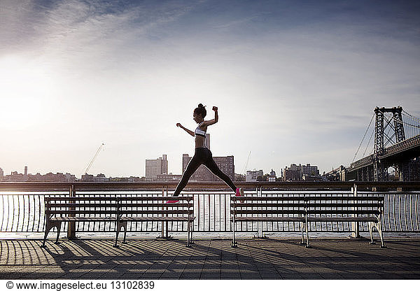 Sporty woman jumping on benches with Williamsburg Bridge in background