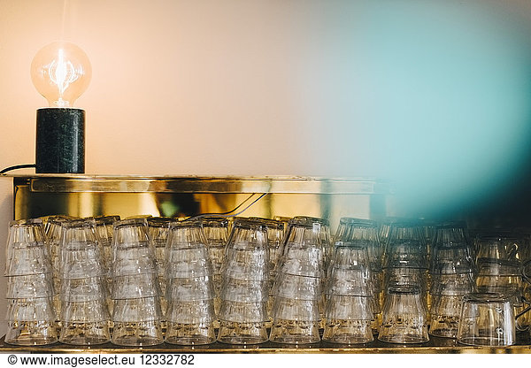 Stack of drinking glasses on shelf by illuminated light bulb against wall in office canteen