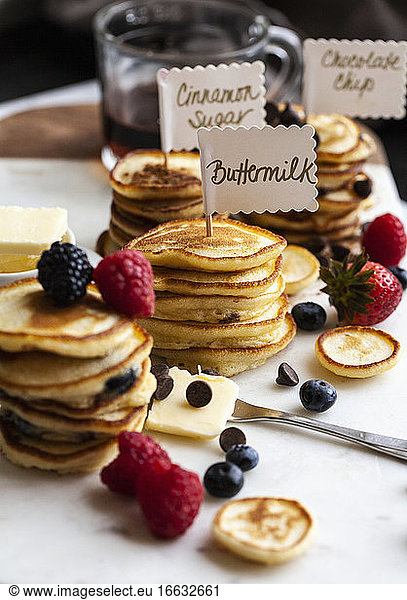 Stacks of mini pancakes with signs  with fruit and chocolate chips  syrup and butter  on a marble board