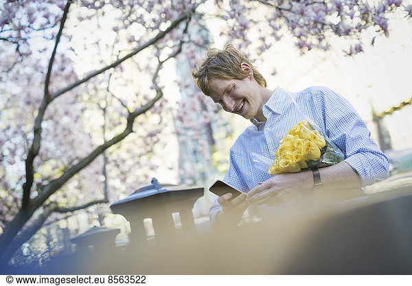 Stadtleben. Ein junger Mann im Frühling im Park  der ein Mobiltelefon benutzt. Er hält einen Strauß gelber Rosen in der Hand.