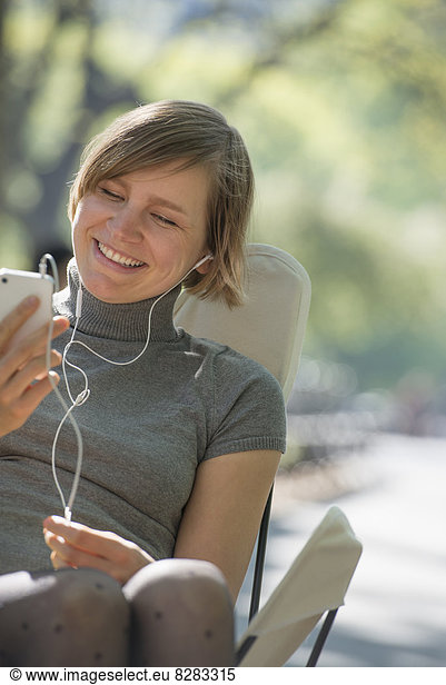Stadtleben. Eine Frau sitzt in einem Campingstuhl im Park und hört mit Kopfhörern Musik.