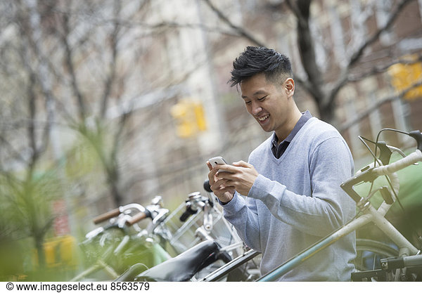 Stadtleben im Frühling. Ein Mann in einem blauen Pullover an einer Reihe geparkter Fahrräder. Seine Nachrichten auf einem Smartphone abrufen.