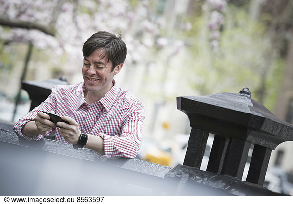 Stadtleben im Frühling. Ein Mann sitzt draußen in einem Stadtpark. Er schaut auf sein Smartphone und lächelt.