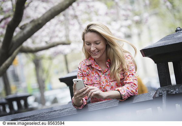 Stadtleben im Frühling. Eine junge Frau mit langen blonden Haaren sitzt in einem Stadtpark und schaut auf ihr Smartphone.
