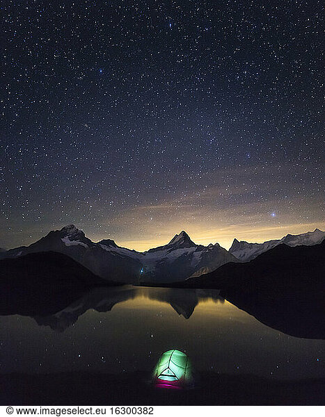 Starry night sky over illuminated tent pitched on shore of Bachalpsee lake at night