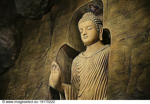 Stehende Buddhastatue in Mönchskleidung  die in eine Kalksteinhöhle gemeißelt und von Licht beleuchtet ist. Rechte Hand in Adhay Mudra Geste. Religion  Tradition  Gebet  Meditation