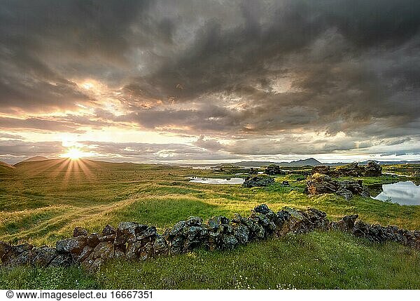 Steinmauer und Lavaformationen aus vulkanischem Gestein ragen aus grüner Wiese mit dramatischem Himmel und Sonnenstern im Abendlicht am Kalfaströnd  See Mývatn  Skútustaðir  Norðurland eystra  Island  Europa