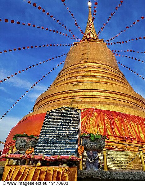 Steintafel mit langer Botschaft in Thai vor einem großen goldenen Stupa oder Chedi  geschmückt mit festlichen roten Fahnen und Textilien im Sonnenuntergang im Wat Saket oder Golden Mount Tempel  Bangkok  Thailand. Traditionelle Opfergaben  Gebet  Anbetung  Botschaften
