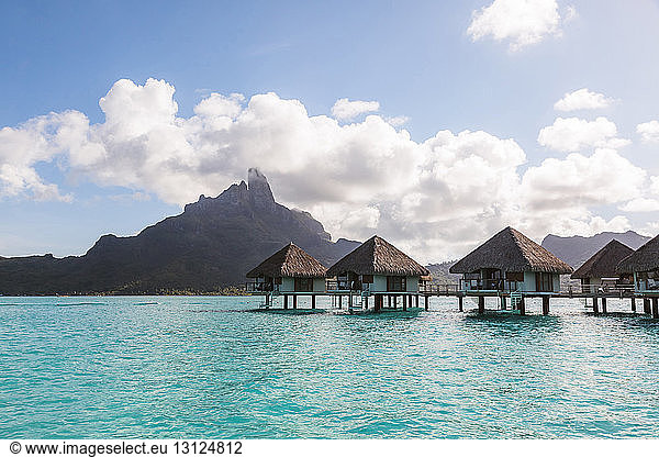 Stilt houses in lagoon of Bora Bora island by mountain against cloudy sky