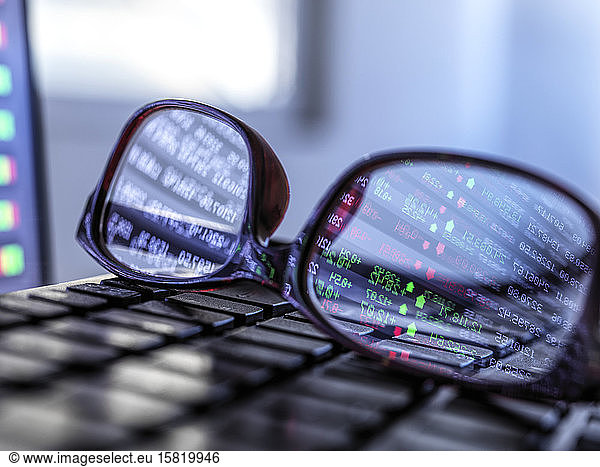 Stock market data reflecting in eyeglasses lying on laptop keyboard