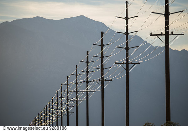 Stromleitungen in Reihen quer durch die Landschaft  vor einem Berg und am Himmel bei Sonnenuntergang.