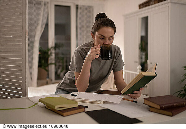 Student reading book and drinking coffee during exam preparation