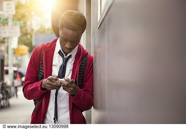 Student using phone while leaning on wall in city