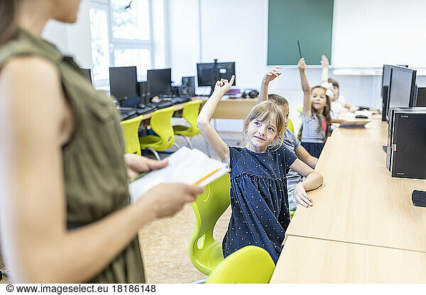 Students with hand raised looking at teacher standing in class at school