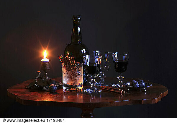 Studio shot of bottle of red wine  three filled wineglasses  plums  salty pretzels and burning candle on small coffee table
