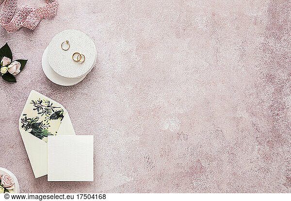 Studio shot of invitation card  ribbon  engagement ring and pair of wedding rings flat laid against pink background