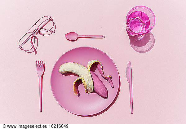 Studioaufnahme von rosa Plastikgeschirr  einem Glas Wasser  einer Brille und einer rosa geschälten Banane