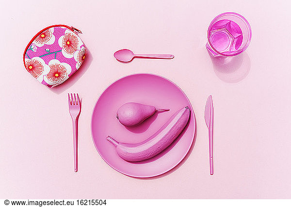 Studioaufnahme von rosa Plastikgeschirr  einem Glas Wasser  rosa Früchten und einer kleinen Tasche mit Blumenmuster