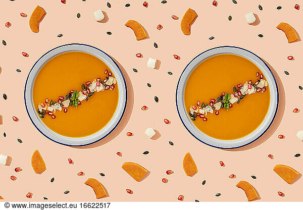 Studioaufnahme von zwei Schalen mit Kürbissuppe  umgeben von Kürbiskernen  Granatapfelkernen und Stücken von Käse und Kürbis