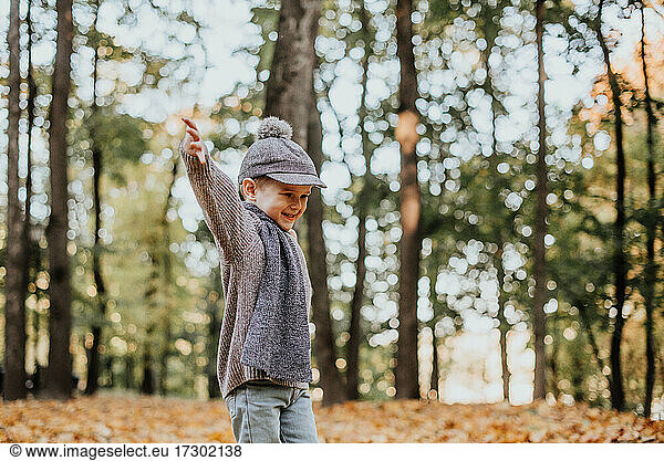 Stylish boy having fun in autumn city park. Happy kid playing woth fallen leaves. Kids fashion. Boy wearing trendy yellow coat  cap and scarf. Smiling young boy outdoors. Kid jumping and runn