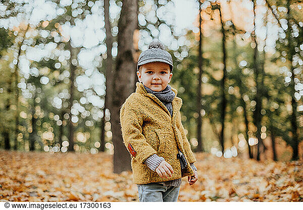 Stylish boy having fun in autumn city park. Happy kid walking among fallen leaves. Kids fashion. Boy wearing trendy yellow coat  cap and scarf. Smiling young boy outdoors. Kid jumping and run