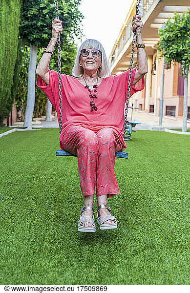 Stylish happy senior woman wearing sunglasses swinging in yard