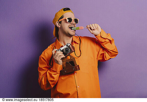 Stylish man in orange shirt with camera and ice cream against purple background