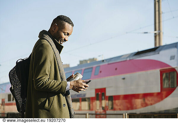 Stylish man with reusable cup using smartphone while waiting for the train