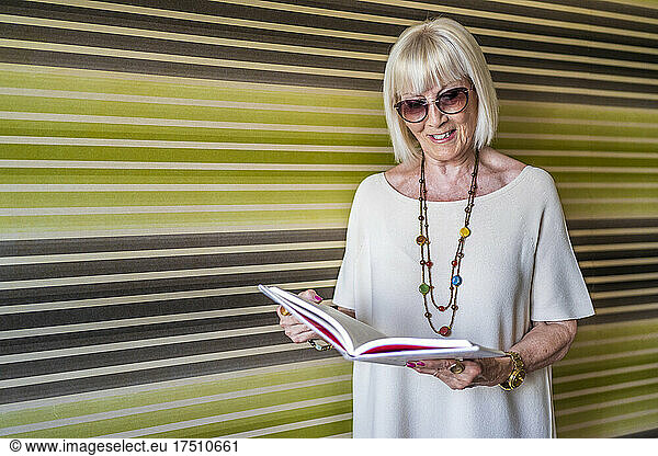 Stylish senior woman wearing sunglasses reading book while standing against wall at home