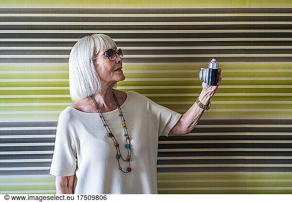 Stylish senior woman wearing sunglasses taking selfie with camera against wall at home