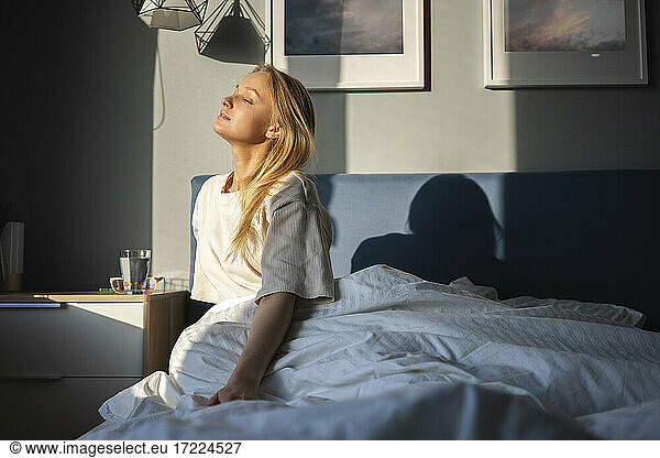 Sunlight on woman sitting with eyes closed on bed at home