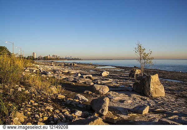 Sunrise on the shore of Lake Michigan. We can see the rocky shore  the lake and the Chicago Skyline. Sky is blue and the sun rising make the horizon of the skyline orange.