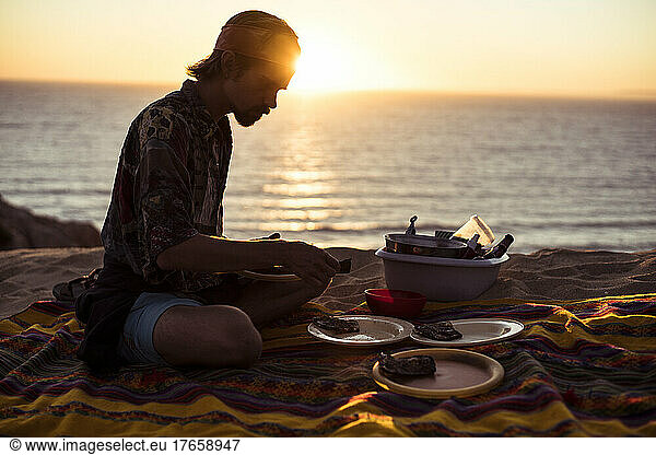 Sunset glow over ocean as man makes picnic dinner in sand Portugal