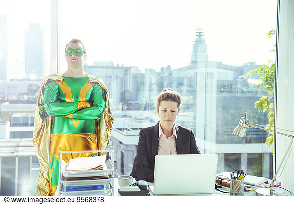 Superhero standing near businesswoman working in office