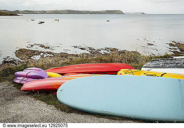 Surf boards and bodyboard  a kayak and boats on the sand on a beach  and view of the coastline and moored boats.