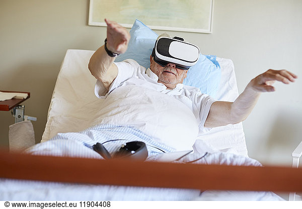 Surface level of senior man with arms raised using VR glasses on bed in hospital ward