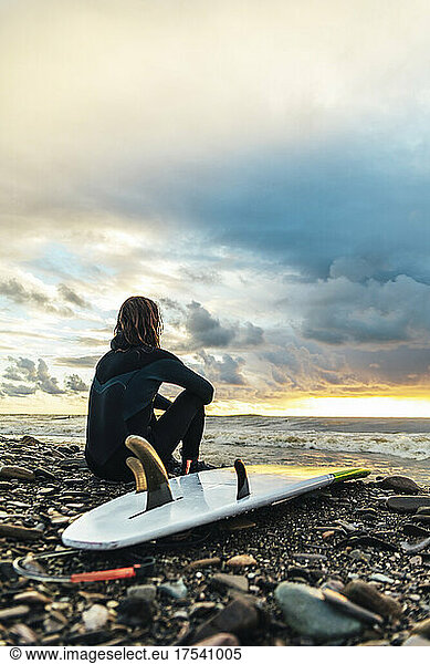 Surfer sitting by surfboard on pebbles looking at sunset over sea