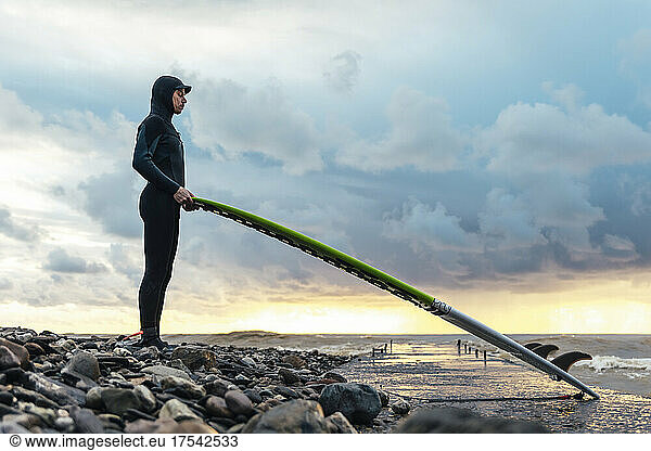 Surfer wearing wetsuit holding surfboard standing on pebbles near sea