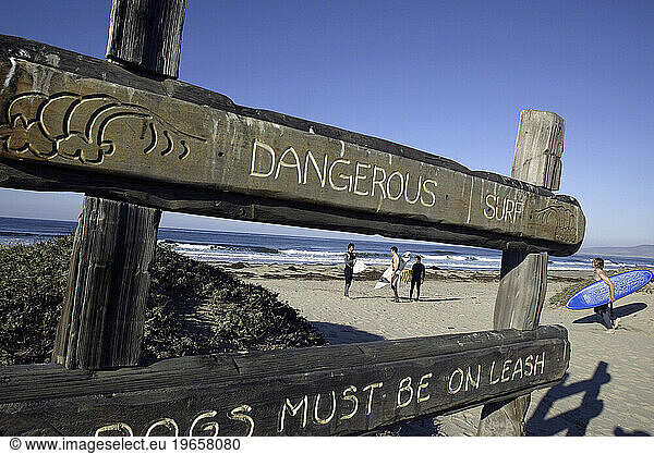 Surfers walking across beach with warning sign.