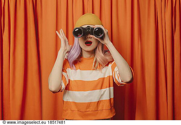 Surprise woman looking through binoculars in front of orange curtain