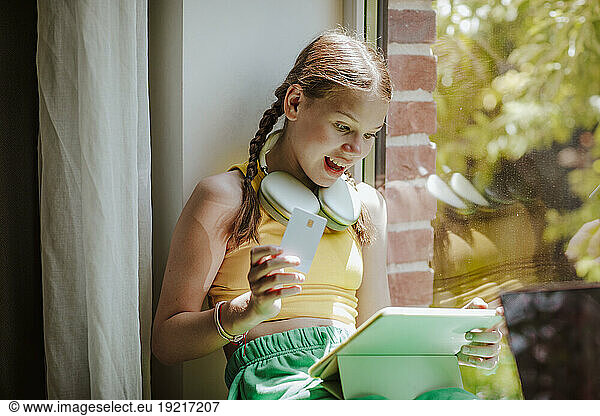 Surprised girl with credit card using tablet PC sitting at home