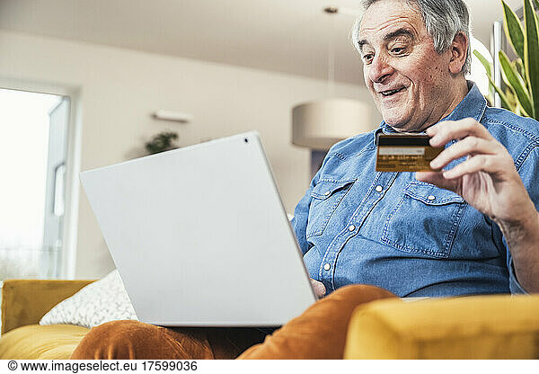 Surprised senior man with credit card using laptop in living room