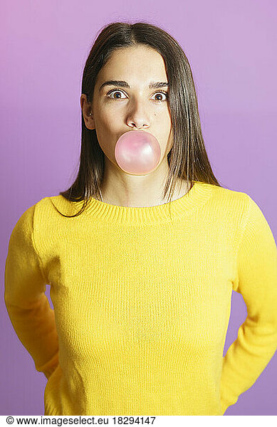 Surprised woman blowing bubble gum standing against purple background