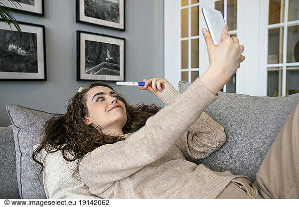 Surprised woman showing pregnancy testing kit on video call at home