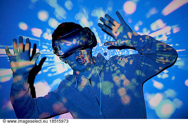 Surprised young man wearing VR glasses standing with hand raised in front of wall