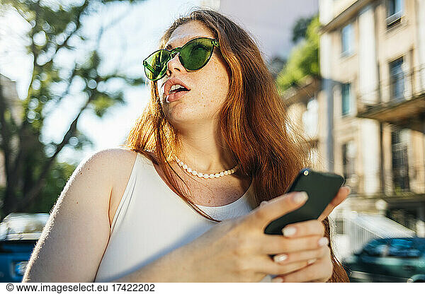 Surprised young woman in green sunglasses holding mobile phone