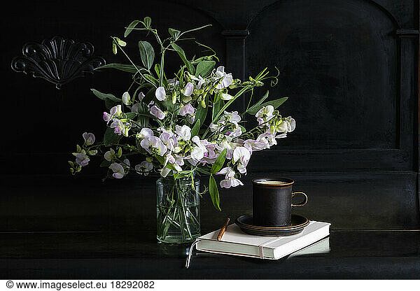 Sweetpea flowers in glass jar by coffee cup and diary on black piano