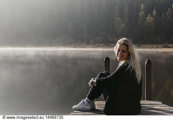 Switzerland, Engadine, Lake Staz, smiling young woman sitting on a jetty at lakeside in morning sun