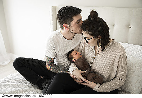 Tätowierter Millennial-Papa küsst Mutter  während sie ihr Neugeborenes im Arm hält