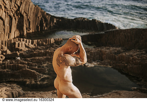 Tätowierter Nudist posiert auf vulkanischen Felsen am Meer